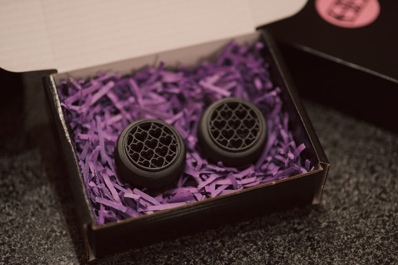 May include: Two black ear gauges with a heart pattern design, displayed in a black box with purple shredded paper.