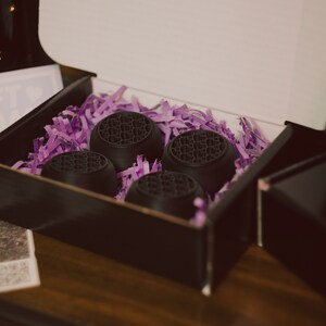 May include: Four black round objects with a textured surface are nestled in purple shredded paper inside a black box. The box is open and the lid is propped up.