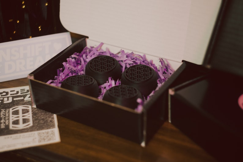 May include: Four black round objects with a textured surface are nestled in purple shredded paper inside a black box. The box is open and the lid is propped up.