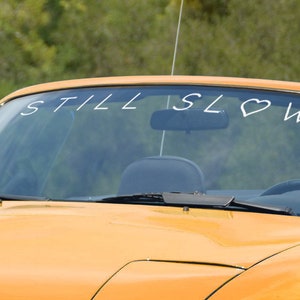 May include: A yellow convertible car with a windshield sticker that reads "STILL SLOW" with a heart symbol in place of the "O" in SLOW.