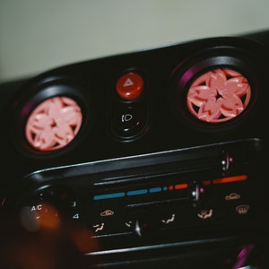 May include: Close-up of a car's dashboard with a black background and pink flower-shaped air freshener vents. The dashboard features a control panel with buttons and knobs, including AC controls and a radio.