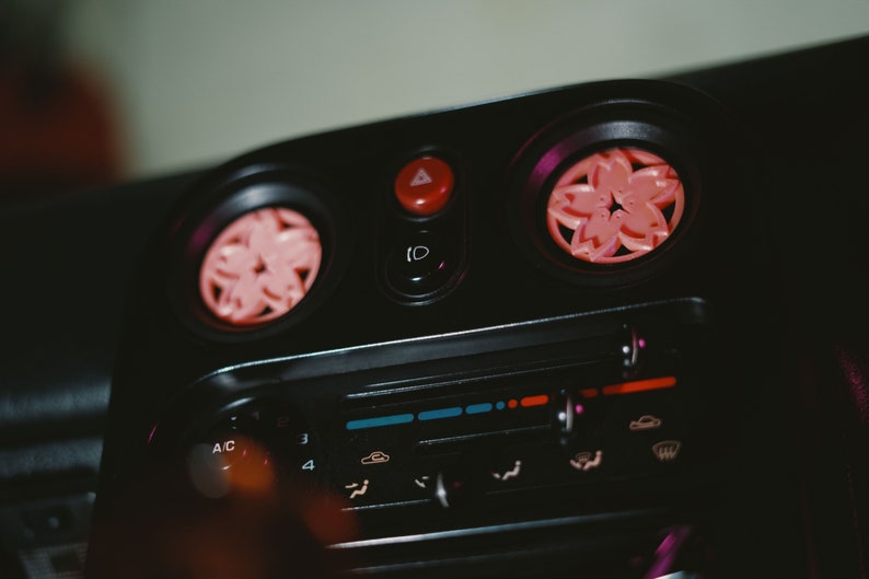 May include: Close-up of a car's dashboard with a black background and pink flower-shaped air freshener vents. The dashboard features a control panel with buttons and knobs, including AC controls and a radio.