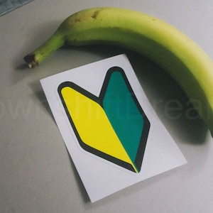 May include: A yellow and green sticker with a black outline in the shape of a Japanese beginner's mark, also known as a "wakaba mark", on a white background. A green banana is lying on a gray surface next to the sticker.