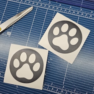 May include: Two square white stickers with a black paw print design, each inside a black circle. The stickers are on a blue cutting mat with a ruler and a pair of scissors.
