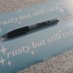 May include: Two light blue stickers with white text and stars that say "rusty but still cute". A silver Sharpie marker is lying on top of the stickers.