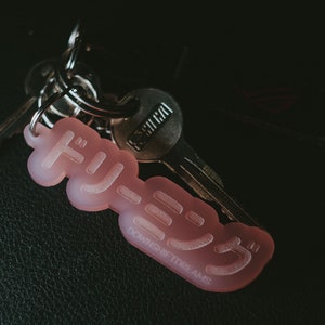 May include: A pink keychain with the Japanese word "Hygge" written on it. The keychain is attached to a silver key ring with a silver key.