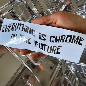 May include: A white sticker with the text "EVERYTHING IS CHROME IN THE FUTURE" in black, held against a shiny, chrome-like surface. The sticker's bold font and reflective background create a striking contrast.