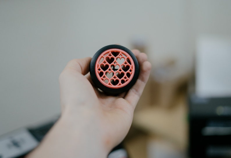 May include: A black and pink 3D printed object with a heart design. The object is being held in a person's hand.