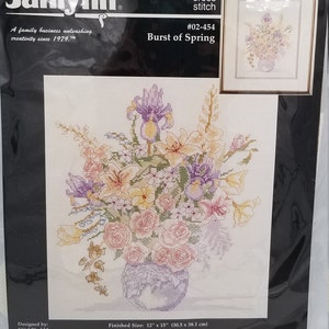 May include: A counted cross stitch pattern featuring a vase of flowers in shades of pink, purple, yellow, and white. The pattern is titled "Burst of Spring" and is from Janlynn, a family business that has been creating cross stitch patterns since 1979. The finished size of the pattern is 12 inches by 15 inches.