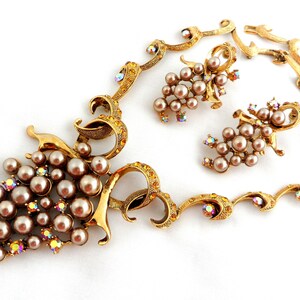 May include: A gold tone necklace and earrings set featuring a grape motif with faux pearls and iridescent rhinestones.