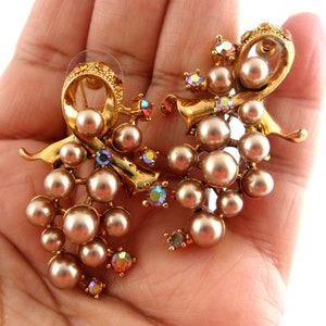 May include: A pair of gold tone earrings with a cluster of faux pearls and iridescent rhinestones. The earrings are shaped like a bow with a cascading cluster of pearls.