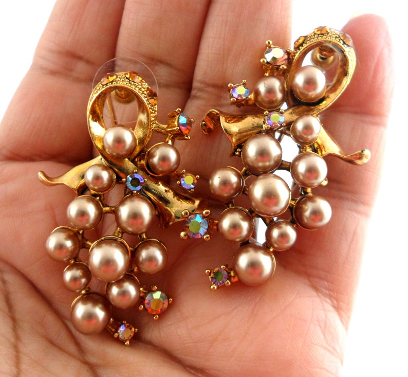 May include: A pair of gold tone earrings with a cluster of faux pearls and iridescent rhinestones. The earrings are shaped like a bow with a cascading cluster of pearls.