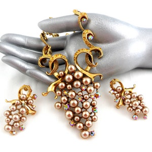 May include: A gold tone necklace and earrings set featuring a grape cluster design with faux pearls and iridescent rhinestones.