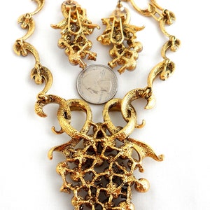 May include: A gold tone necklace and earrings set with a large, ornate pendant featuring a grapevine design with clear rhinestones. The necklace is made of a delicate chain with a similar grapevine design.
