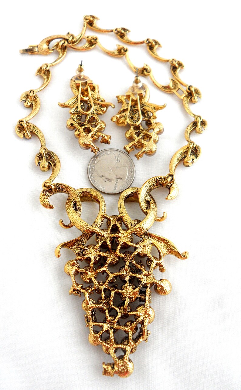 May include: A gold tone necklace and earrings set with a large, ornate pendant featuring a grapevine design with clear rhinestones. The necklace is made of a delicate chain with a similar grapevine design.