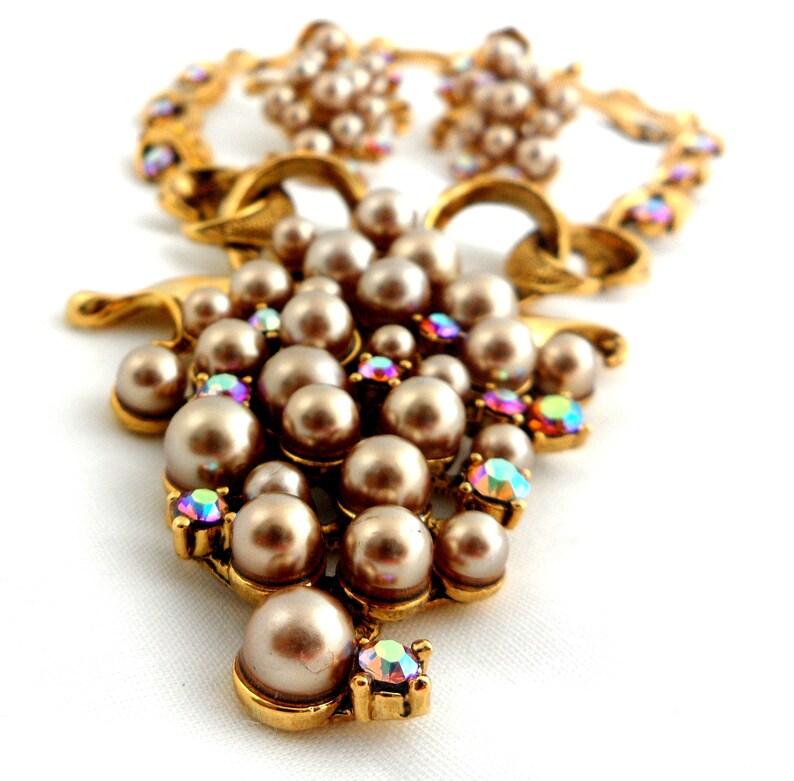 May include: A gold tone necklace and earrings set with a cluster of faux pearls and iridescent rhinestones. The necklace features a large cluster of pearls and rhinestones in the shape of a grape.