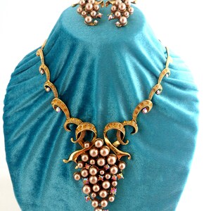 May include: A gold tone necklace and earrings set with a grape cluster design. The necklace features a large cluster of faux pearls and rhinestones, while the earrings feature smaller clusters of faux pearls and rhinestones.