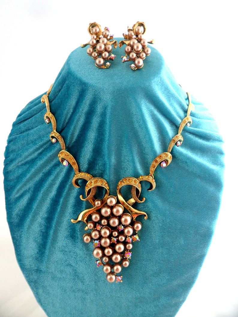 May include: A gold tone necklace and earrings set with a grape cluster design. The necklace features a large cluster of faux pearls and rhinestones, while the earrings feature smaller clusters of faux pearls and rhinestones.