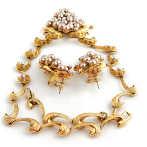 May include: A gold tone necklace and earrings set with a floral design. The necklace has a clasp closure and the earrings are clip-on. The set is adorned with faux pearls and iridescent rhinestones.