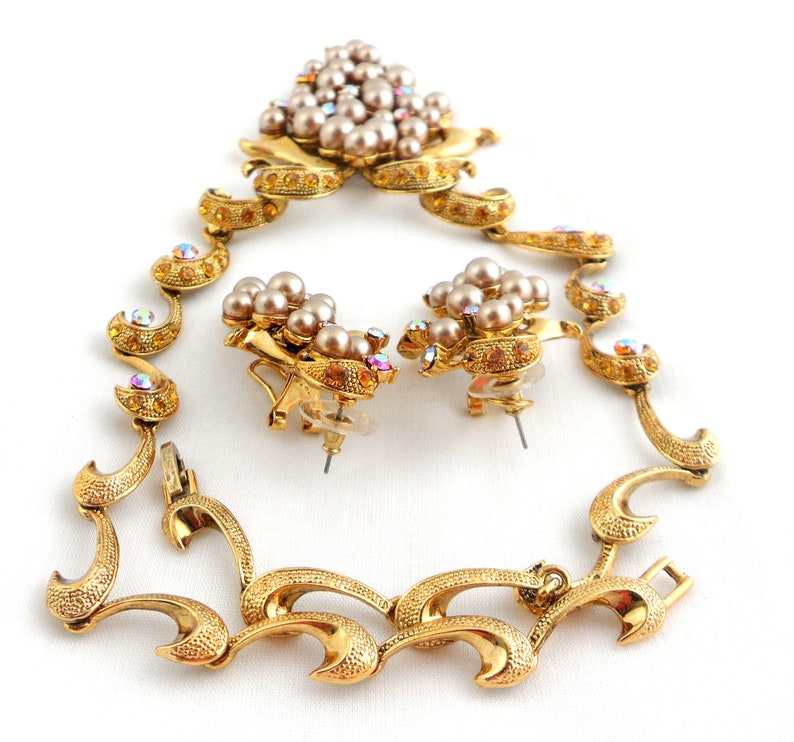 May include: A gold tone necklace and earrings set with a floral design. The necklace has a clasp closure and the earrings are clip-on. The set is adorned with faux pearls and iridescent rhinestones.