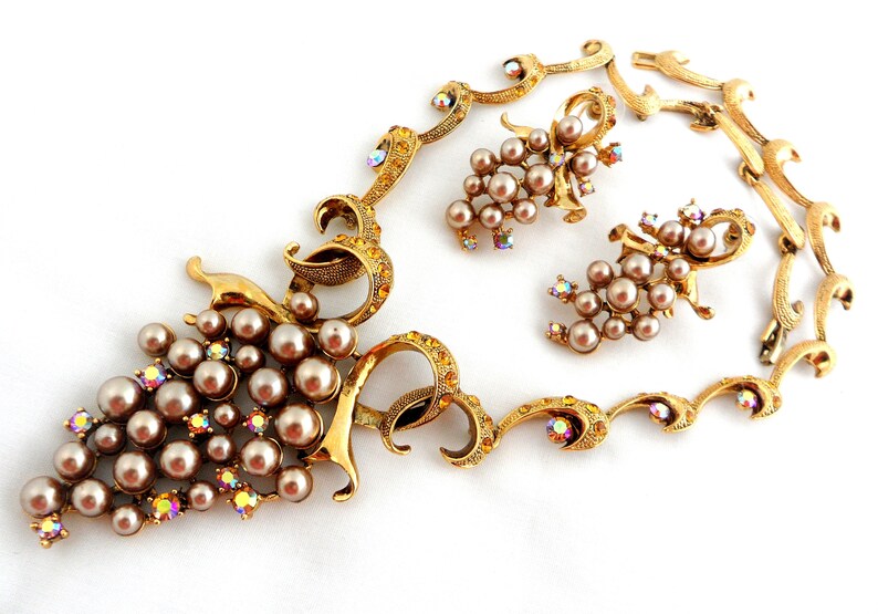 May include: A gold tone necklace and earrings set featuring a grape motif with faux pearls and iridescent rhinestones.