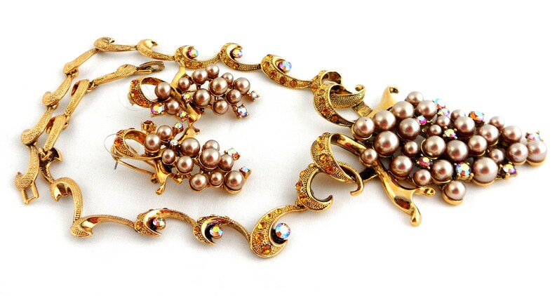 May include: A gold-tone necklace and earrings set featuring a grape motif with faux pearls and iridescent rhinestones.