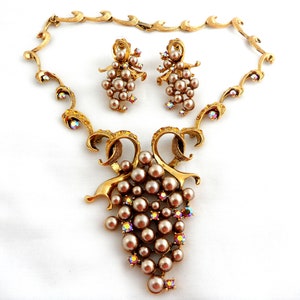 May include: A gold tone necklace and earrings set with a grape cluster design. The necklace features a delicate chain with a pendant of a grape cluster made of faux pearls and iridescent rhinestones. The earrings are drop earrings with a similar grape cluster design.