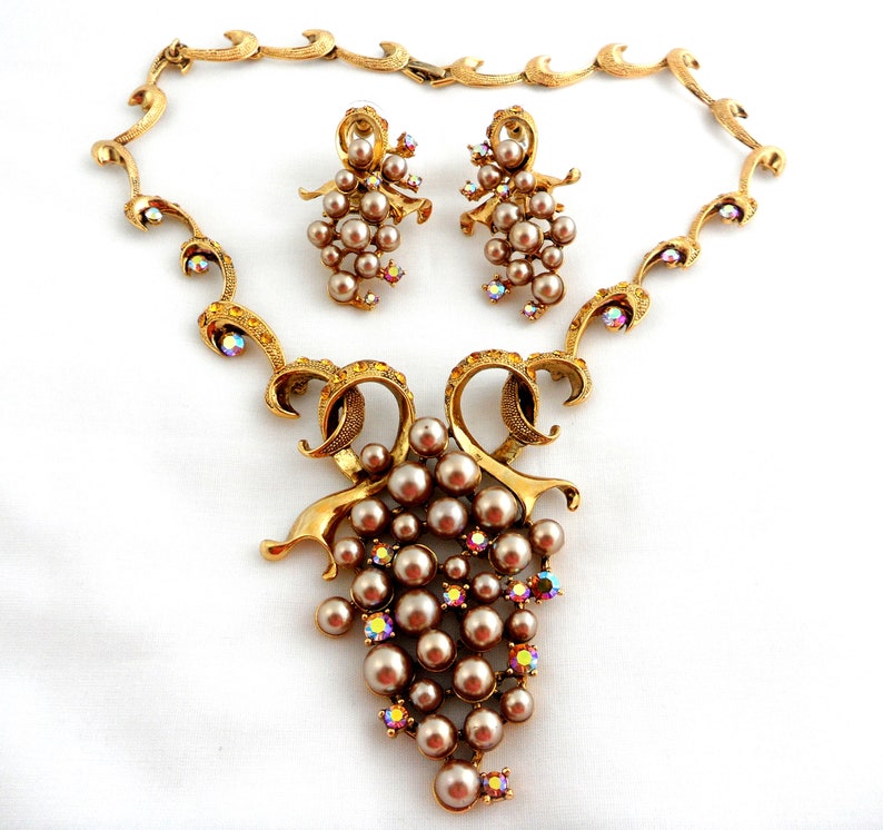 May include: A gold tone necklace and earrings set with a grape cluster design. The necklace features a delicate chain with a pendant of a grape cluster made of faux pearls and iridescent rhinestones. The earrings are drop earrings with a similar grape cluster design.