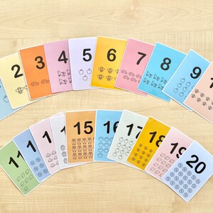 Puede incluir: Un conjunto de veinte tarjetas de memoria coloridas con los n&uacute;meros del 1 al 20. Cada tarjeta tiene un color diferente y presenta un n&uacute;mero correspondiente de peque&ntilde;os iconos, como manzanas, abejas, coches y b&uacute;hos.