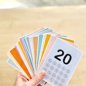 Puede incluir: Un conjunto de 20 tarjetas coloridas con un dise&ntilde;o de baloncesto y el n&uacute;mero 20 impreso en el frente. Las tarjetas est&aacute;n abiertas en abanico en una mano.