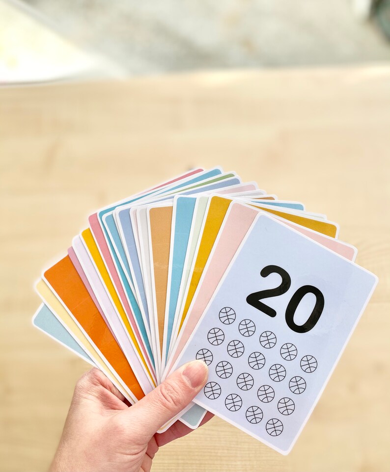 Puede incluir: Un conjunto de 20 tarjetas coloridas con un dise&ntilde;o de baloncesto y el n&uacute;mero 20 impreso en el frente. Las tarjetas est&aacute;n abiertas en abanico en una mano.