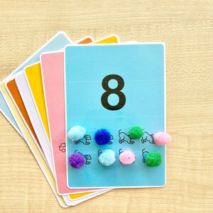 Puede incluir: Una tarjeta azul con el n&uacute;mero 8 impreso en ella. Hay ocho pompones de colores en la tarjeta, cada uno colocado junto a un dibujo en blanco y negro de un perro.