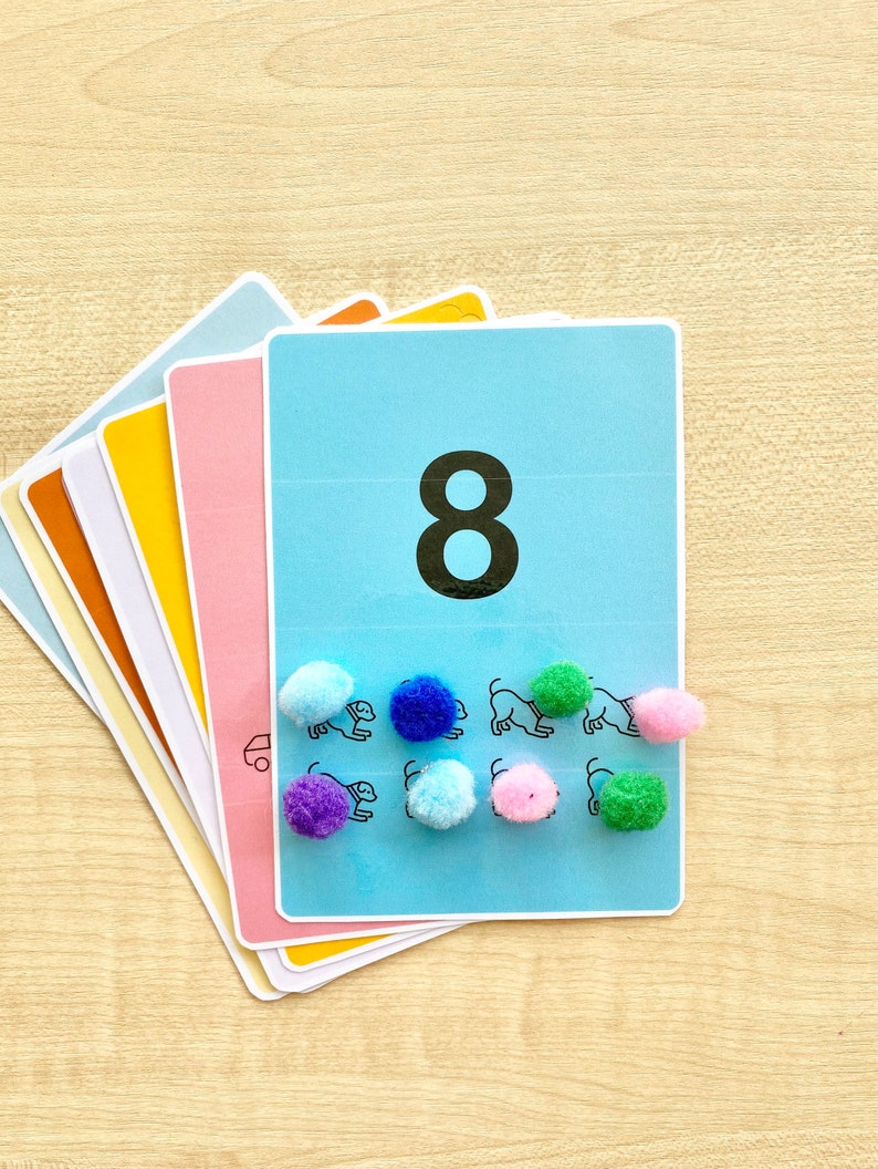 Puede incluir: Una tarjeta azul con el n&uacute;mero 8 impreso en ella. Hay ocho pompones de colores en la tarjeta, cada uno colocado junto a un dibujo en blanco y negro de un perro.