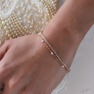 Peut inclure: Un bracelet en or orne un poignet. Il est orn&eacute; de petites pierres transparentes. Une pochette blanche perl&eacute;e avec un fermoir est tenue dans la main. Une bague en or avec une pierre carr&eacute;e est &eacute;galement visible.
