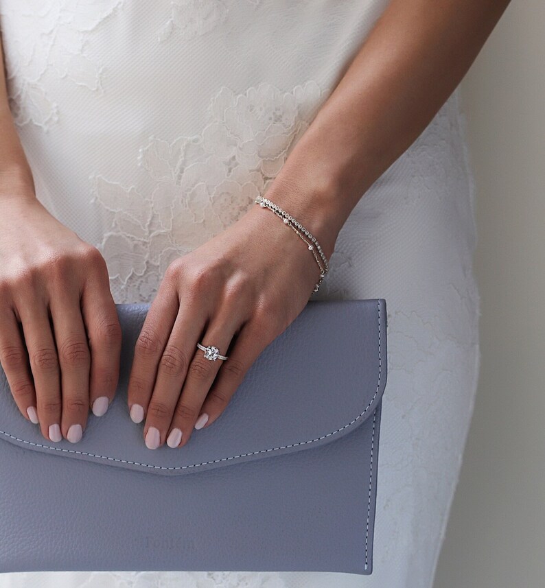 Peut inclure: Une bague de fian&ccedil;ailles en argent avec une grosse pierre pr&eacute;cieuse ronde et transparente est pr&eacute;sent&eacute;e sur une main. Un bracelet d&eacute;licat avec de petites pierres transparentes est sur le poignet. Une pochette bleu clair est tenue dans l'autre main. Le fond est une robe blanche.