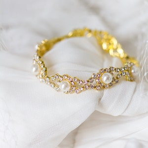 May include: A gold bracelet with a delicate design featuring small white pearls and clear rhinestones. The bracelet is lying on a white fabric background.