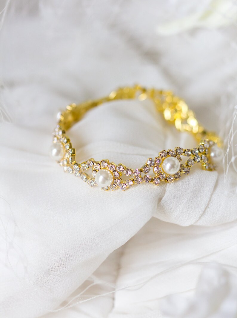 May include: A gold bracelet with a delicate design featuring small white pearls and clear rhinestones. The bracelet is lying on a white fabric background.