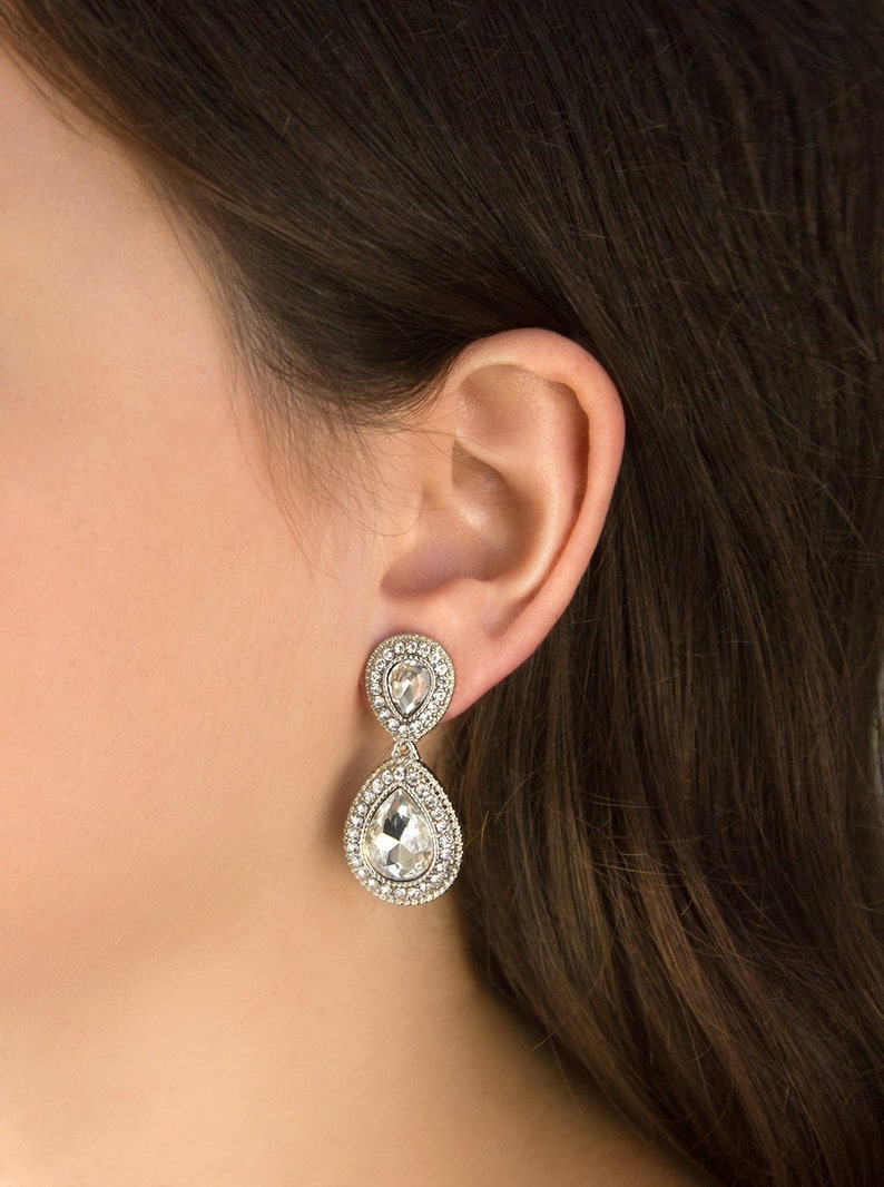 May include: A pair of silver teardrop earrings with clear crystal stones. The earrings have a double teardrop design with smaller stones surrounding the larger stones.