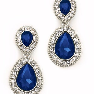 May include: A pair of dangle earrings with a teardrop-shaped blue gemstone surrounded by a halo of clear rhinestones. The earrings have a silver tone finish.