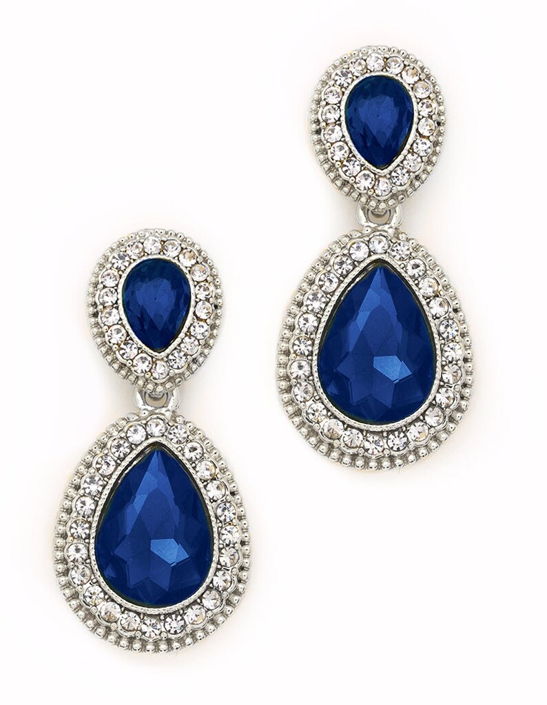 May include: A pair of dangle earrings with a teardrop-shaped blue gemstone surrounded by a halo of clear rhinestones. The earrings have a silver tone finish.