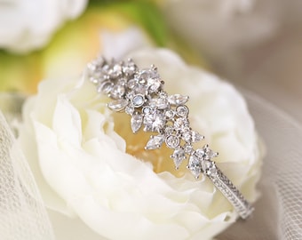 May include: A silver bracelet with a floral design featuring clear rhinestones. The bracelet is resting on a white flower.