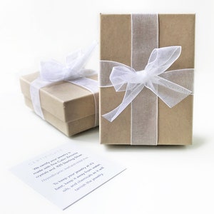 Puede incluir: Dos cajas de regalo marrones con cinta y lazos blancos. Las cajas est&aacute;n sobre una superficie blanca. Una tarjeta blanca con el texto "CERTIFICATE" y un mensaje sobre la joyer&iacute;a est&aacute; en primer plano.