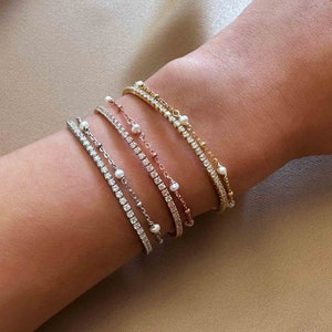 Peut inclure: Plusieurs bracelets d&eacute;licats en or, argent et or rose. Les bracelets sont orn&eacute;s de pierres claires scintillantes et de petites perles blanches. Les bracelets sont superpos&eacute;s sur un poignet.