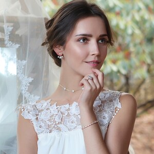 Peut inclure: Une femme porte une robe blanche et un ensemble de bijoux floraux. L'ensemble comprend des boucles d'oreilles, un collier, une bague et un bracelet. Les bijoux pr&eacute;sentent de petits motifs floraux d&eacute;licats. La robe a une superposition de dentelle.