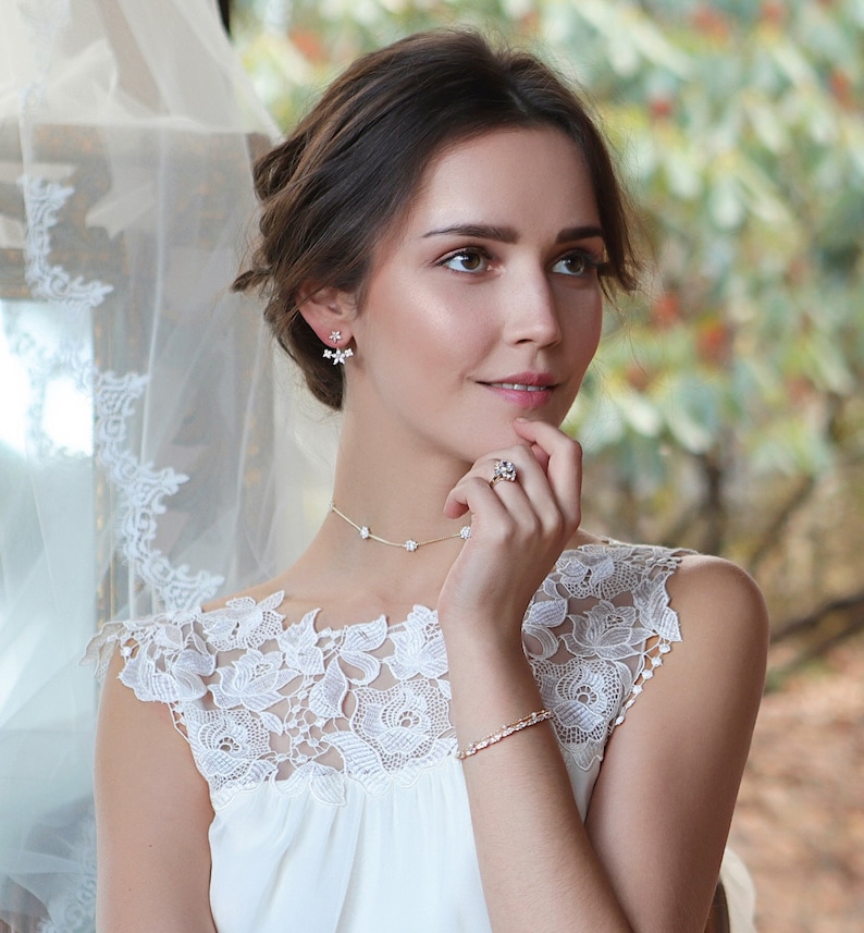 Peut inclure: Une femme porte une robe blanche et un ensemble de bijoux floraux. L'ensemble comprend des boucles d'oreilles, un collier, une bague et un bracelet. Les bijoux pr&eacute;sentent de petits motifs floraux d&eacute;licats. La robe a une superposition de dentelle.