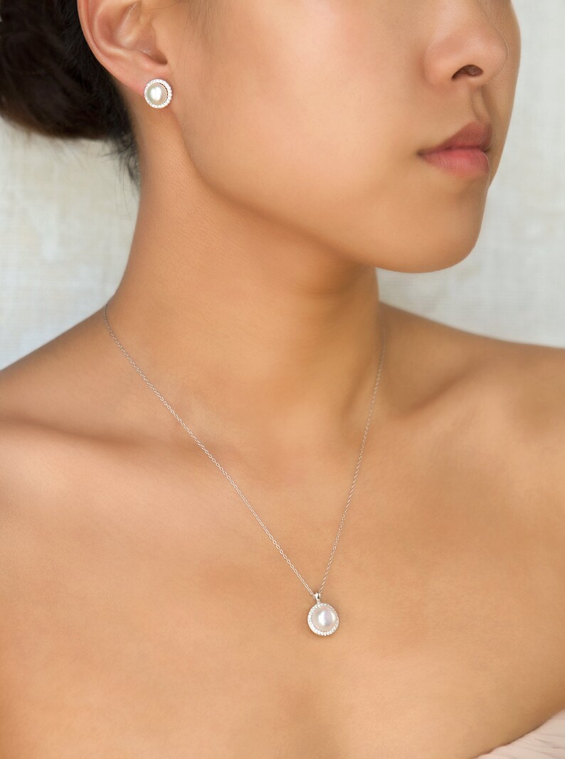 Puede incluir: Un collar de plata con un colgante de perla rodeado de peque&ntilde;os diamantes. El colgante es redondo y cuelga de una cadena delicada. El collar lo lleva una persona de piel clara.