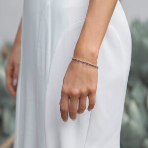 Peut inclure: Un bracelet d&eacute;licat orn&eacute; de petites pierres scintillantes est port&eacute; au poignet. Le bracelet est de couleur argent&eacute;e et semble &ecirc;tre compos&eacute; de plusieurs brins. La personne porte une robe blanche.