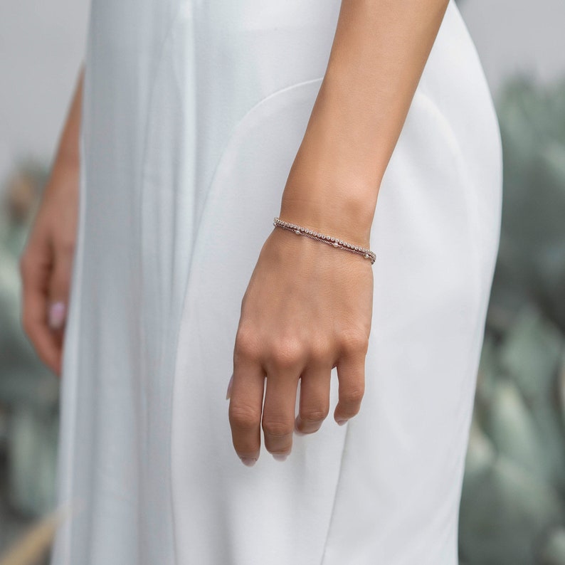 Peut inclure: Un bracelet d&eacute;licat orn&eacute; de petites pierres scintillantes est port&eacute; au poignet. Le bracelet est de couleur argent&eacute;e et semble &ecirc;tre compos&eacute; de plusieurs brins. La personne porte une robe blanche.