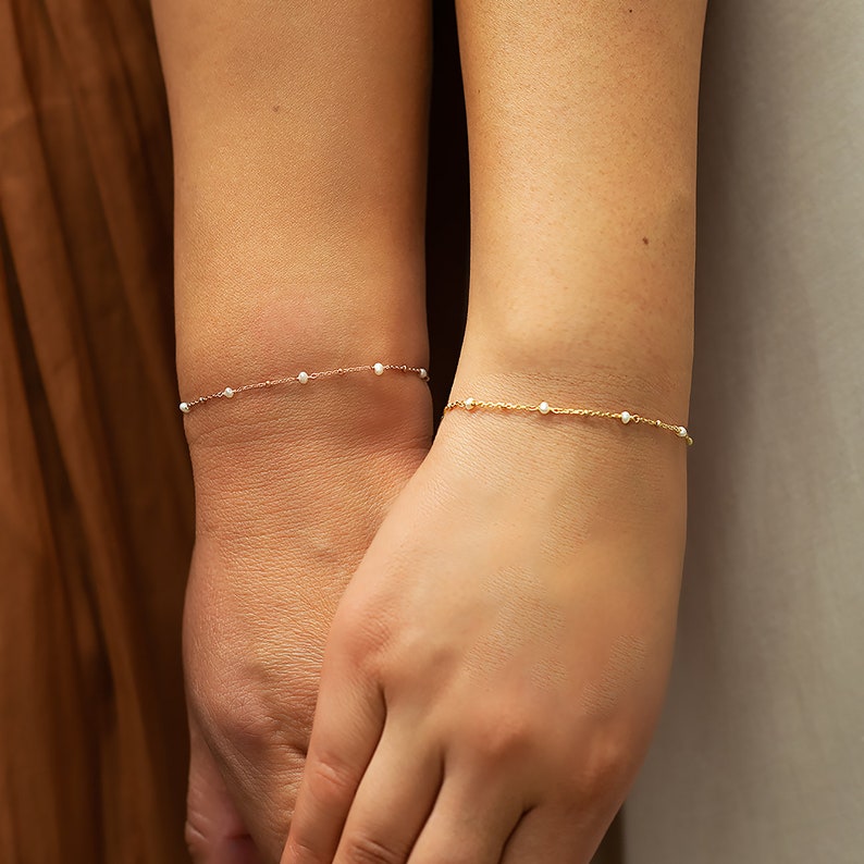 Peut inclure: Deux bracelets fins, l'un en or rose et l'autre en or, chacun orn&eacute; de petites perles blanches. Les bracelets sont port&eacute;s aux poignets, mettant en valeur leur &eacute;l&eacute;gance simple. Le fond est neutre, soulignant les bijoux.