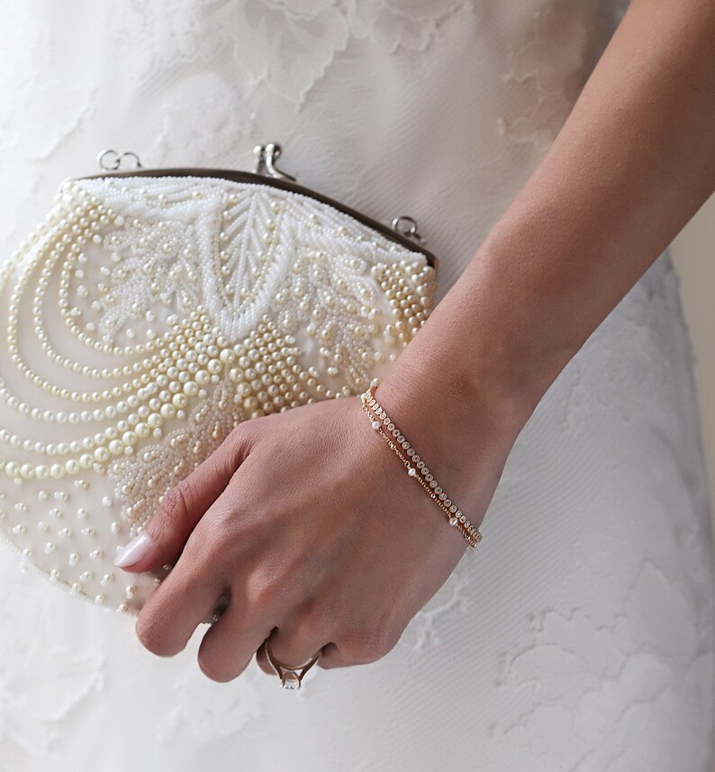Peut inclure: Un bracelet en or orne un poignet. Il est orn&eacute; de petites pierres transparentes. Une pochette blanche perl&eacute;e avec un fermoir est tenue dans la main. Une bague en or avec une pierre carr&eacute;e est &eacute;galement visible.